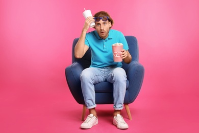 Photo of Emotional man with 3D glasses, popcorn and beverage sitting in armchair during cinema show on color background