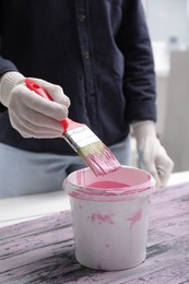 Photo of Woman dipping brush into bucket of pink paint indoors, closeup