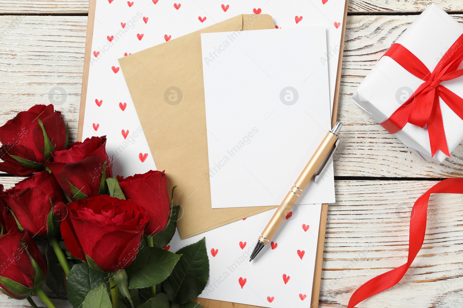 Photo of Flat lay composition with blank greeting card, gift and roses on white wooden table. Valentine's day celebration