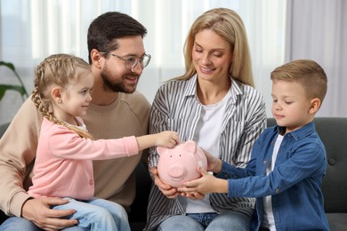 Photo of Family budget. Little girl putting coin into piggy bank while her parents and brother watching indoors