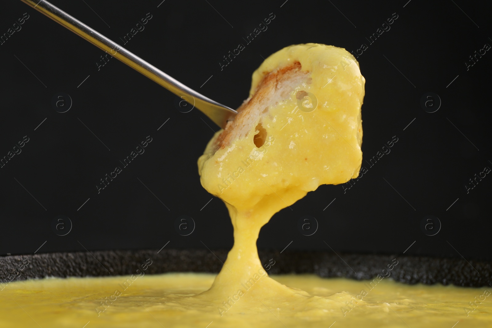 Photo of Dipping piece of bread into fondue pot with tasty melted cheese against dark gray background, closeup