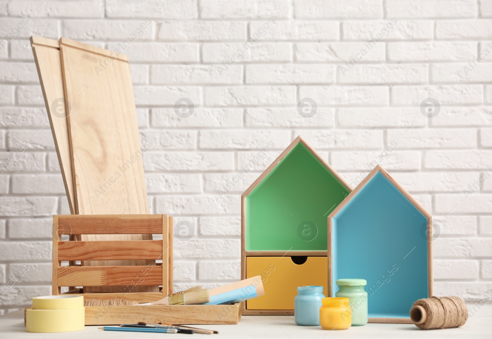 Photo of Composition with house shaped shelves and jars of paints on table against brick wall. Interior elements