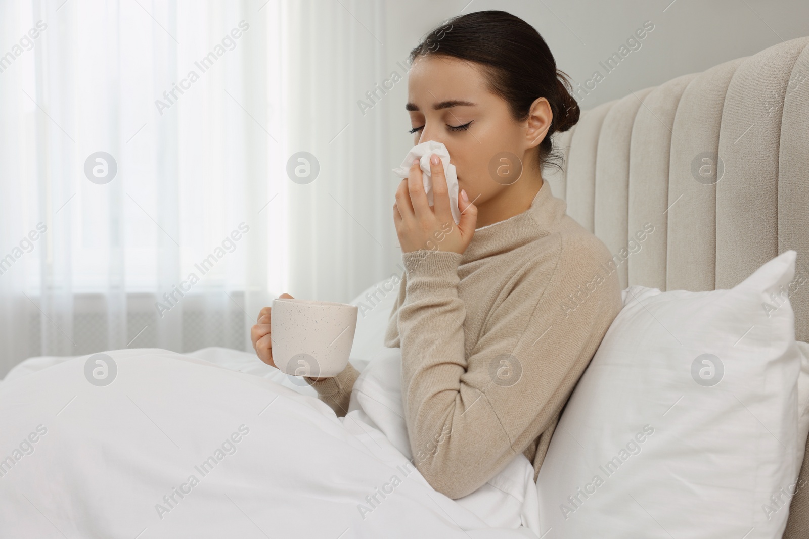 Photo of Sick young woman with cup of hot drink and tissue in bed at home, space for text