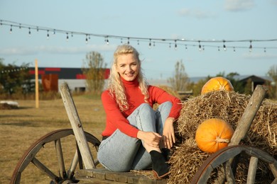 Photo of Beautiful woman sitting on wooden cart with pumpkins and hay in field. Autumn season