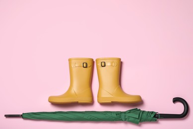 Photo of Pair of yellow rubber boots near green umbrella on pink background, flat lay. Space for text