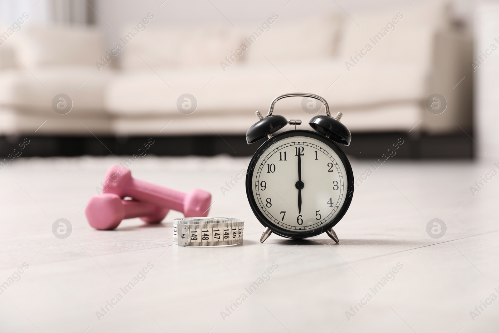 Photo of Alarm clock, measuring tape and dumbbells on wooden floor indoors. Morning exercise