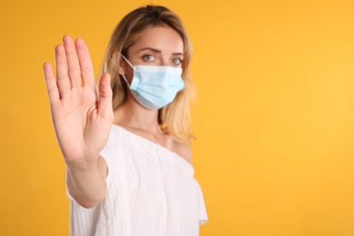 Photo of Woman in protective face mask showing stop gesture on yellow background, focus on hand. Prevent spreading of coronavirus
