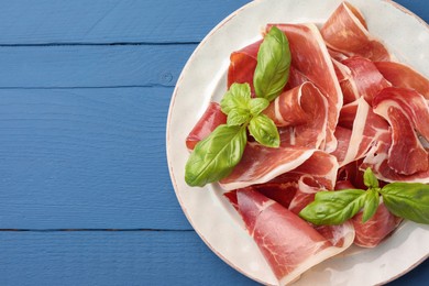 Photo of Slices of tasty cured ham and basil on blue wooden table, top view. Space for text