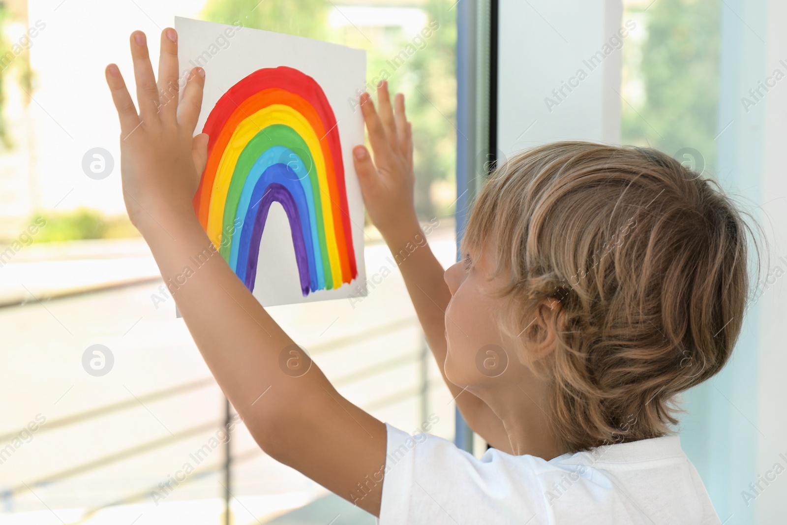 Photo of Little boy holding rainbow painting near window indoors. Stay at home concept