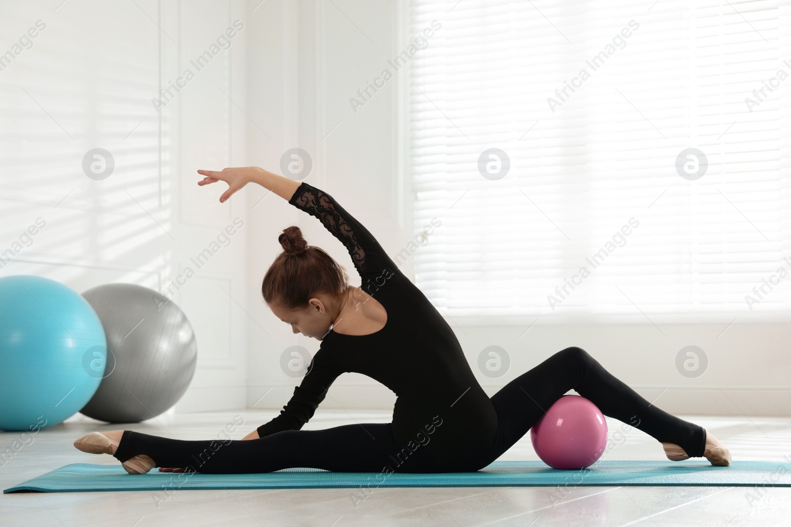 Photo of Cute little gymnast with ball stretching on mat indoors