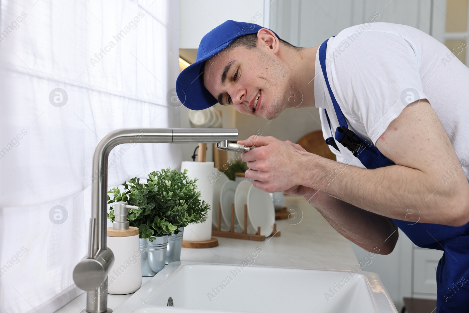 Photo of Smiling plumber repairing faucet with spanner in kitchen