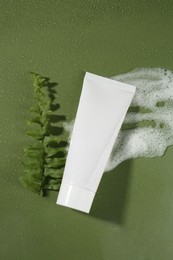 Cleansing foam, tube of cosmetic product and branch on wet dark green background, flat lay