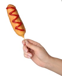 Woman holding delicious deep fried corn dog with ketchup on white background, closeup