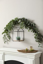 Beautiful garland made of eucalyptus branches and aromatic candle hanging above mantelpiece indoors