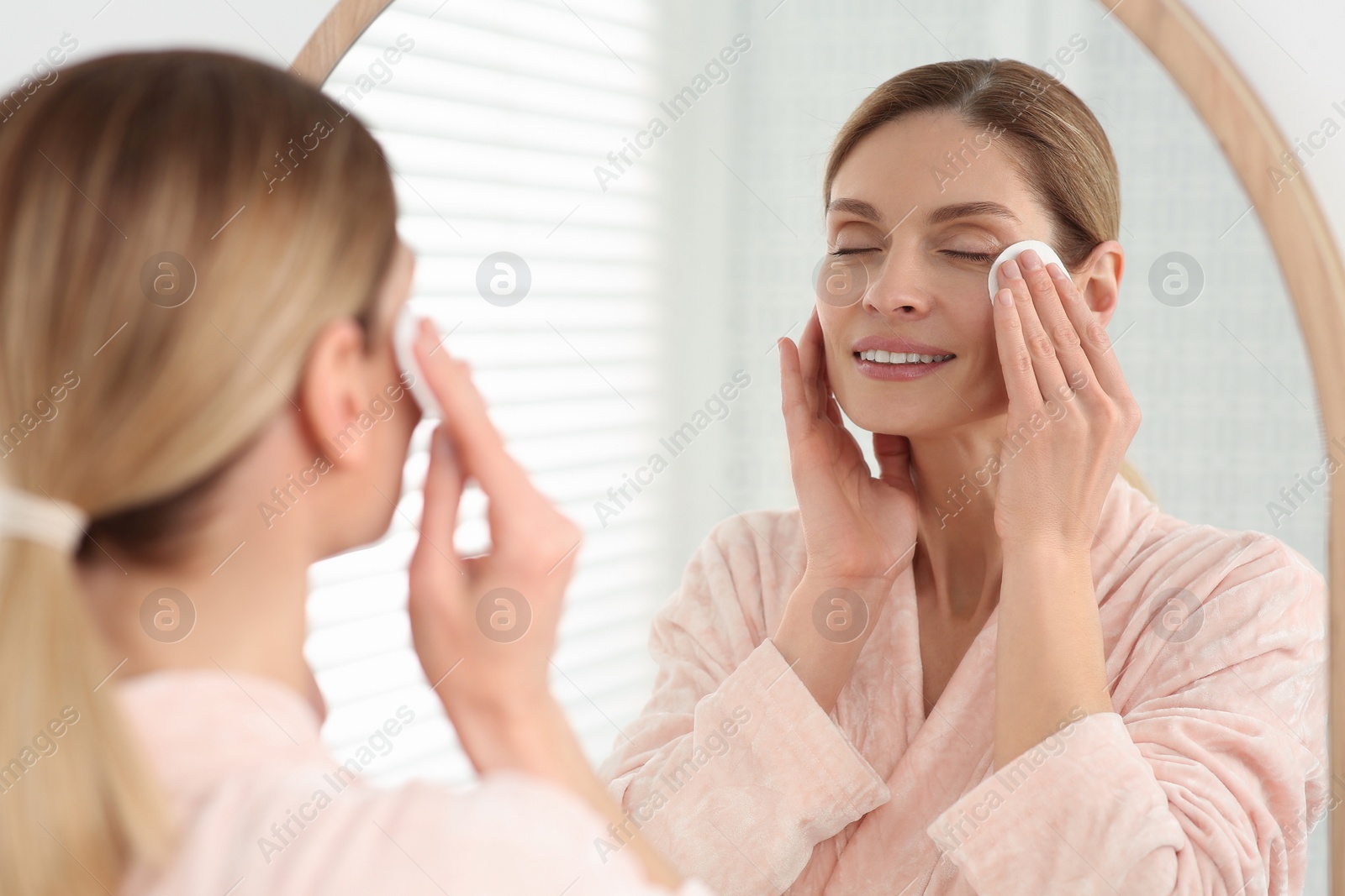 Photo of Beautiful woman removing makeup with cotton pad near mirror indoors