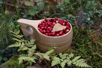 Photo of Many tasty ripe lingonberries and leaf in wooden cup near fern outdoors