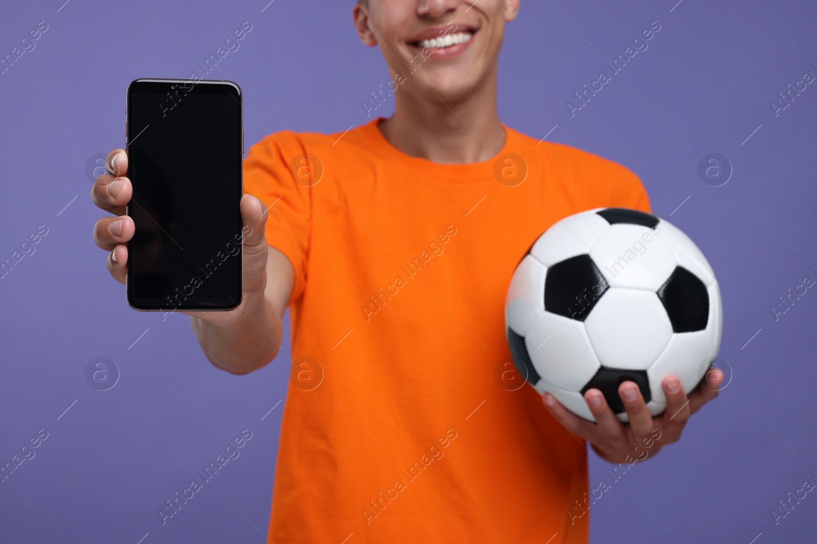Photo of Happy sports fan with soccer ball showing smartphone on purple background, closeup