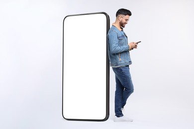 Image of Man with mobile phone standing near huge device with empty screen on grey background. Mockup for design