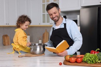 Photo of Cute little girl and her father with recipe book cooking in kitchen