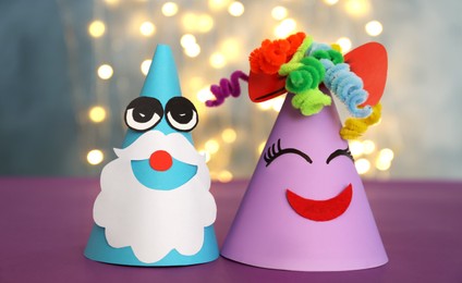 Photo of Funny handmade party hats on purple table against blurred lights, closeup