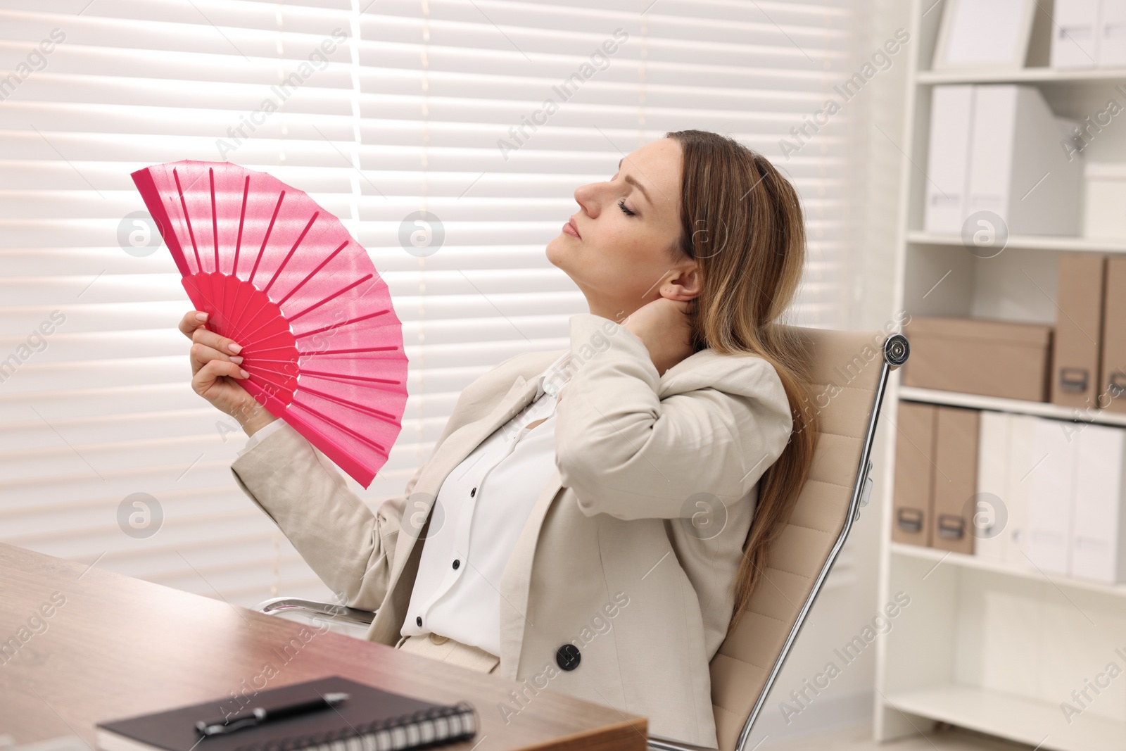 Photo of Businesswoman waving pink hand fan to cool herself at table in office