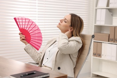 Businesswoman waving pink hand fan to cool herself at table in office