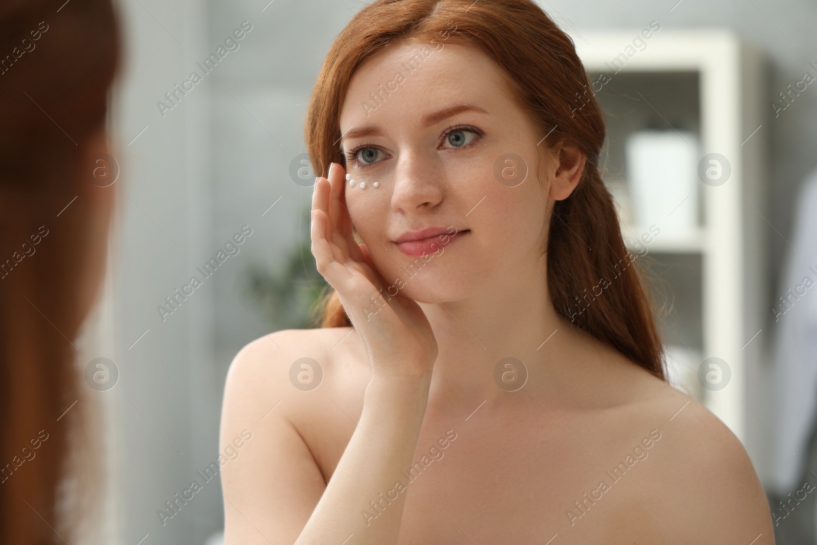 Photo of Beautiful woman with freckles and cream on her face near mirror in bathroom