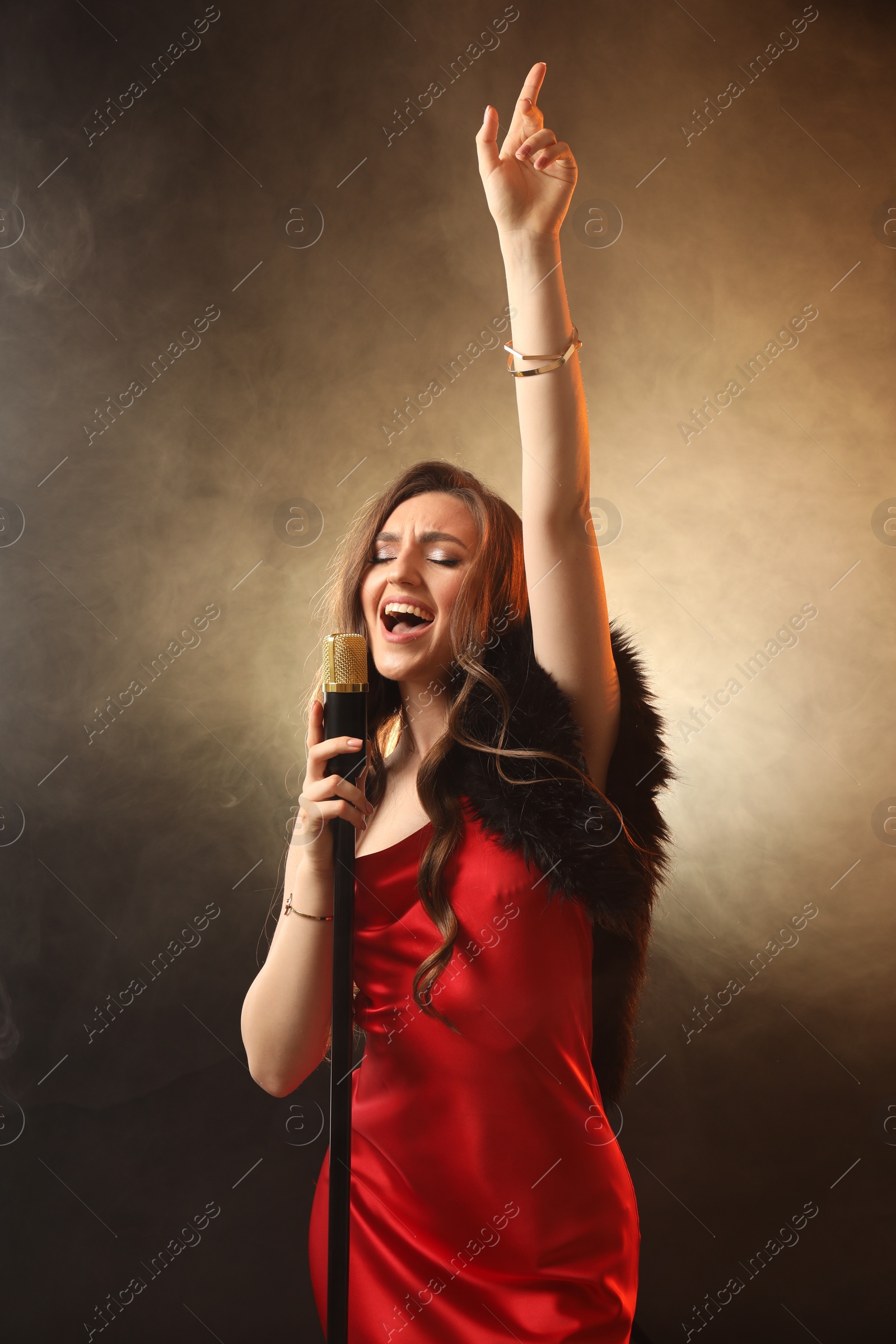 Photo of Beautiful young woman in stylish dress with microphone singing on dark background in color lights and smoke