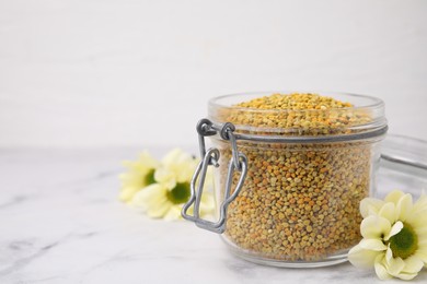 Photo of Fresh bee pollen granules in jar and flowers on light marble table, closeup. Space for text
