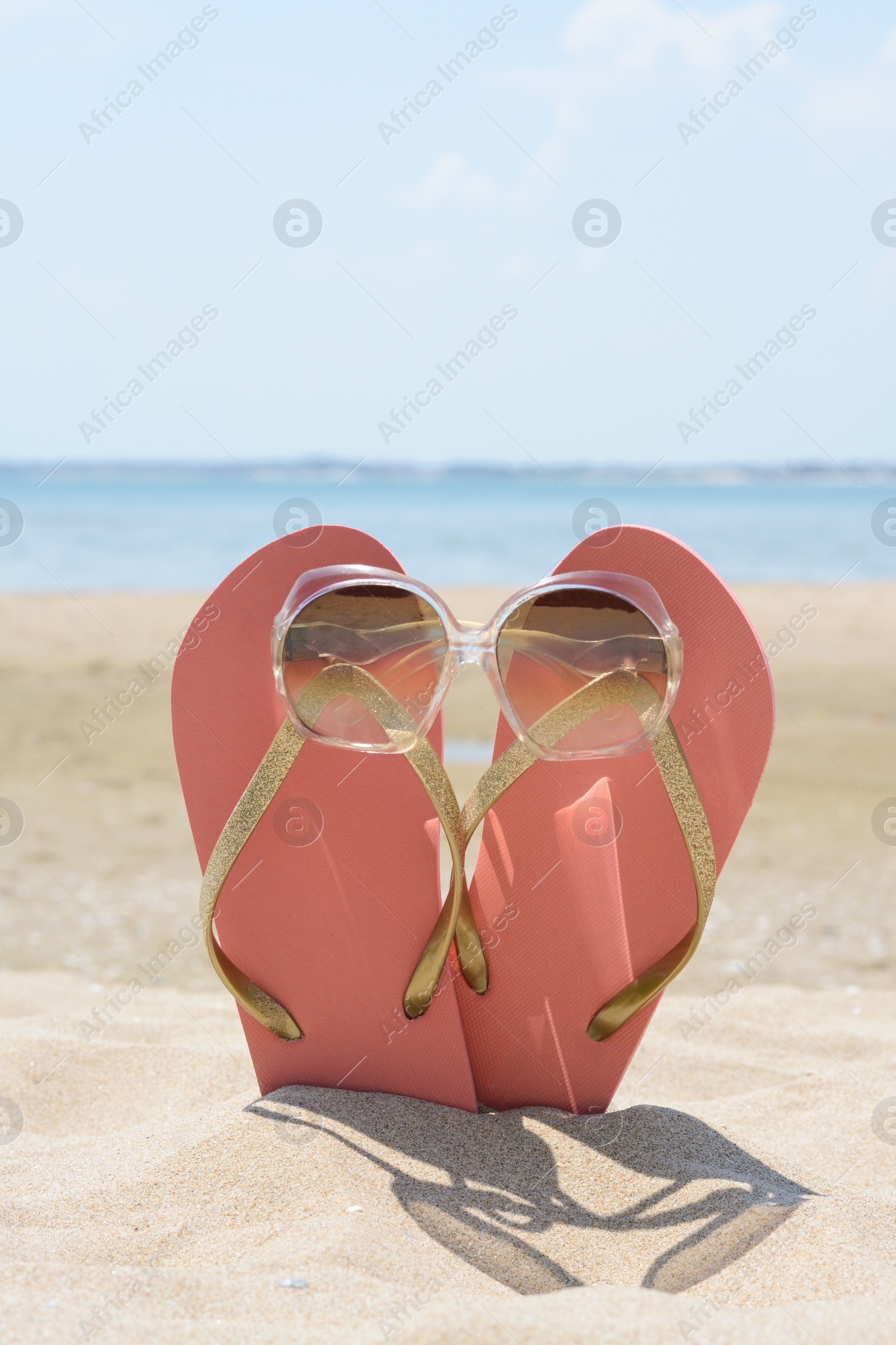 Photo of Stylish pink flip flops with sunglasses in sand near sea on sunny day