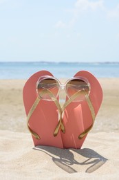 Stylish pink flip flops with sunglasses in sand near sea on sunny day