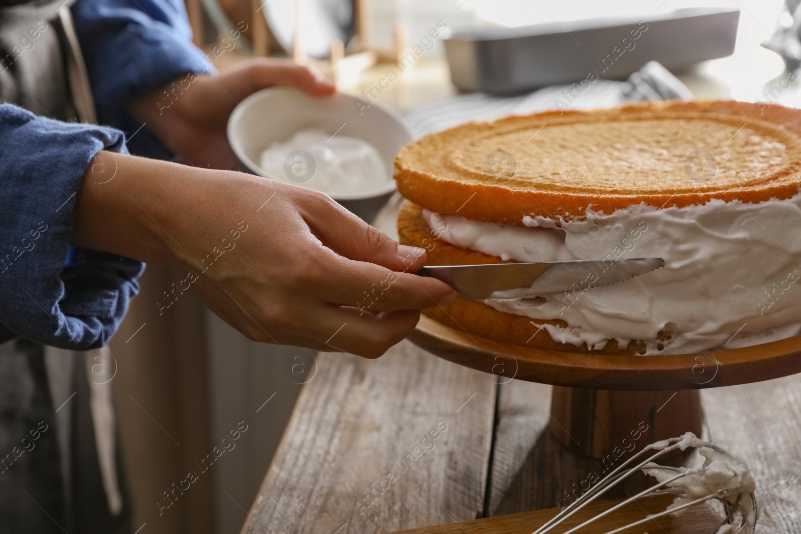 Photo of Woman smearing sides of sponge cake with cream at wooden table, closeup