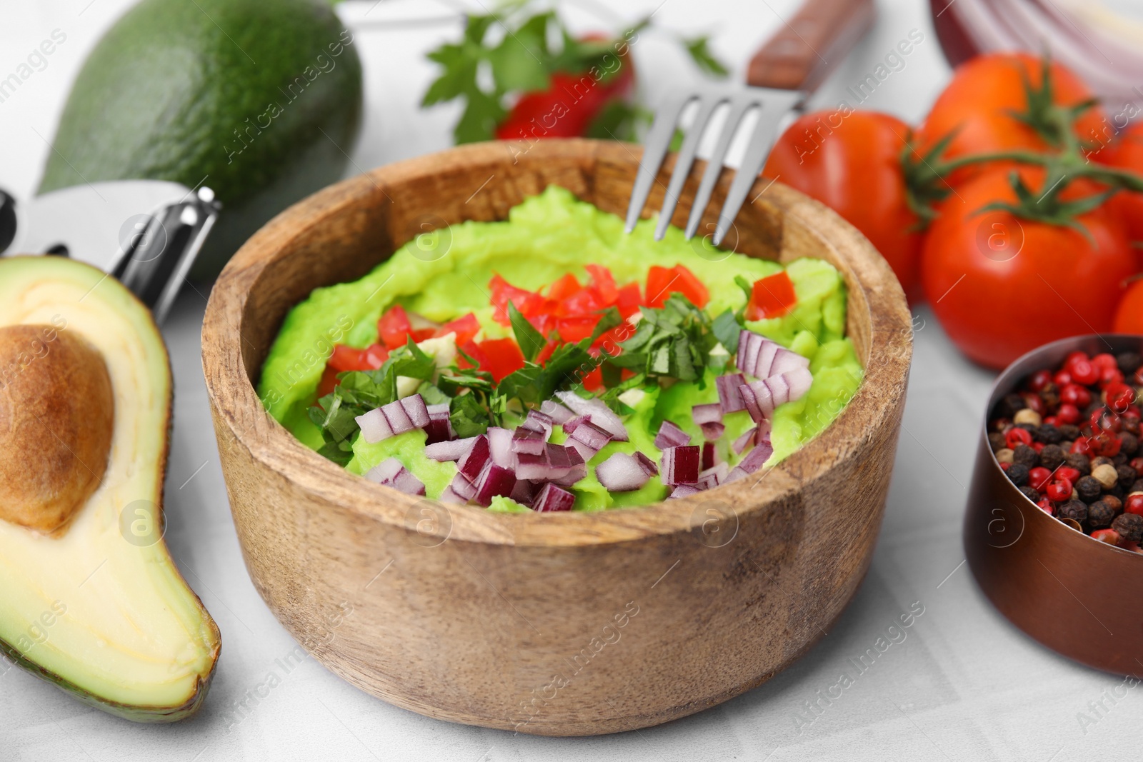 Photo of Bowl of delicious guacamole with onion, tomatoes and ingredients on white tiled table, closeup