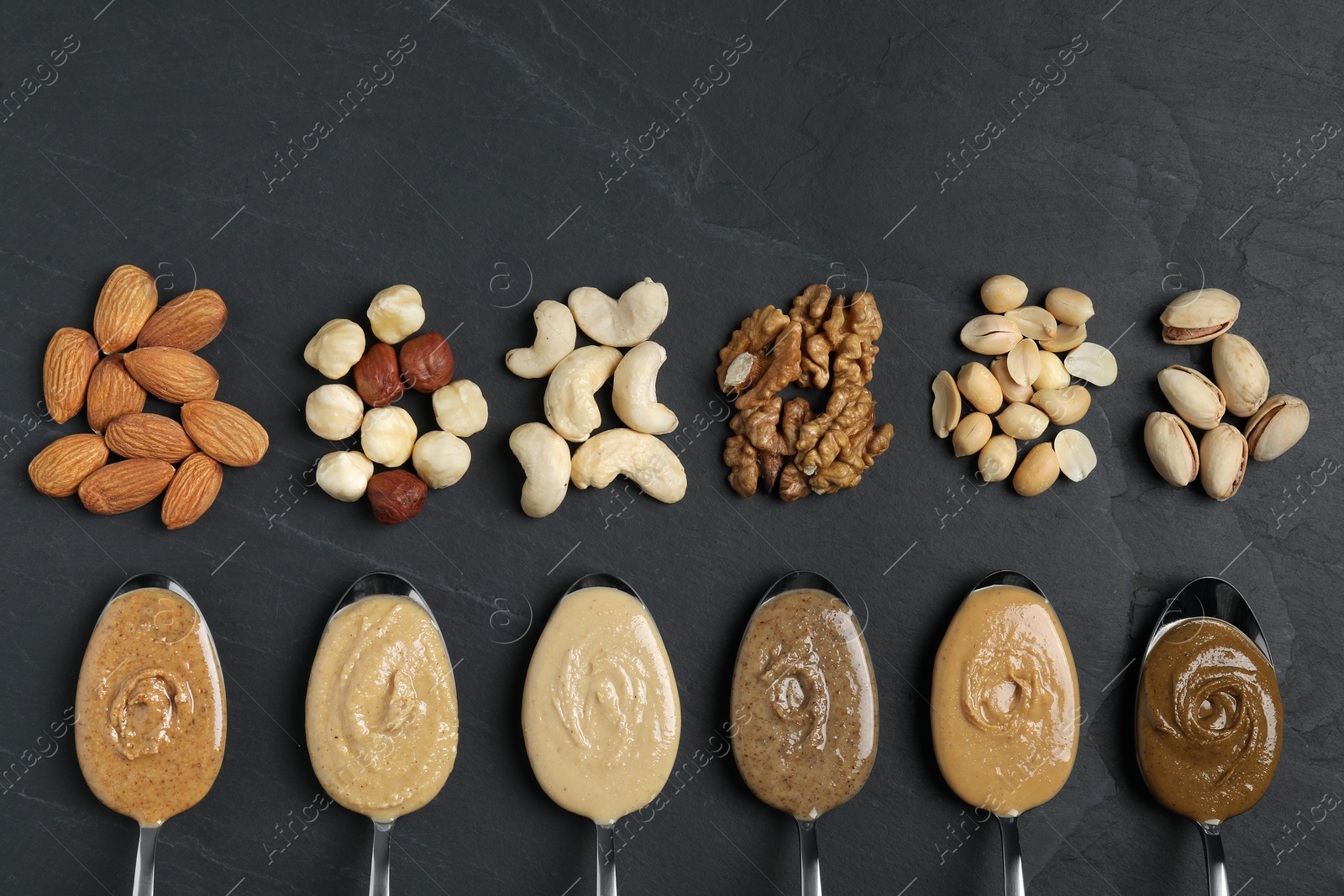Photo of Tasty nut butters in spoons and raw nuts on dark gray textured table, flat lay. Space for text
