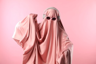 Photo of Glamorous ghost. Woman in sheet with sunglasses and headphones on pink background