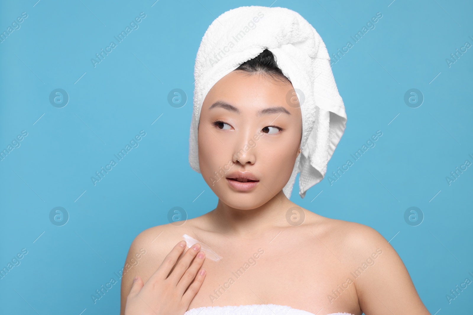 Photo of Beautiful young Asian woman applying body cream onto shoulder on light blue background, space for text