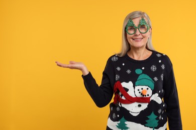 Photo of Happy senior woman in Christmas sweater and funny glasses showing something on orange background. Space for text
