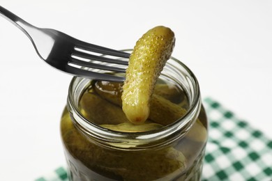 Photo of Fork with pickled cucumber over jar on white table, closeup