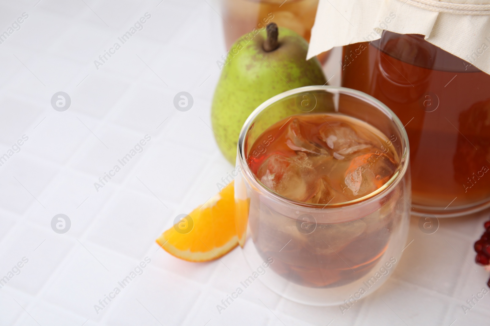 Photo of Tasty kombucha with ice cubes and fresh fruits on white tiled table, space for text