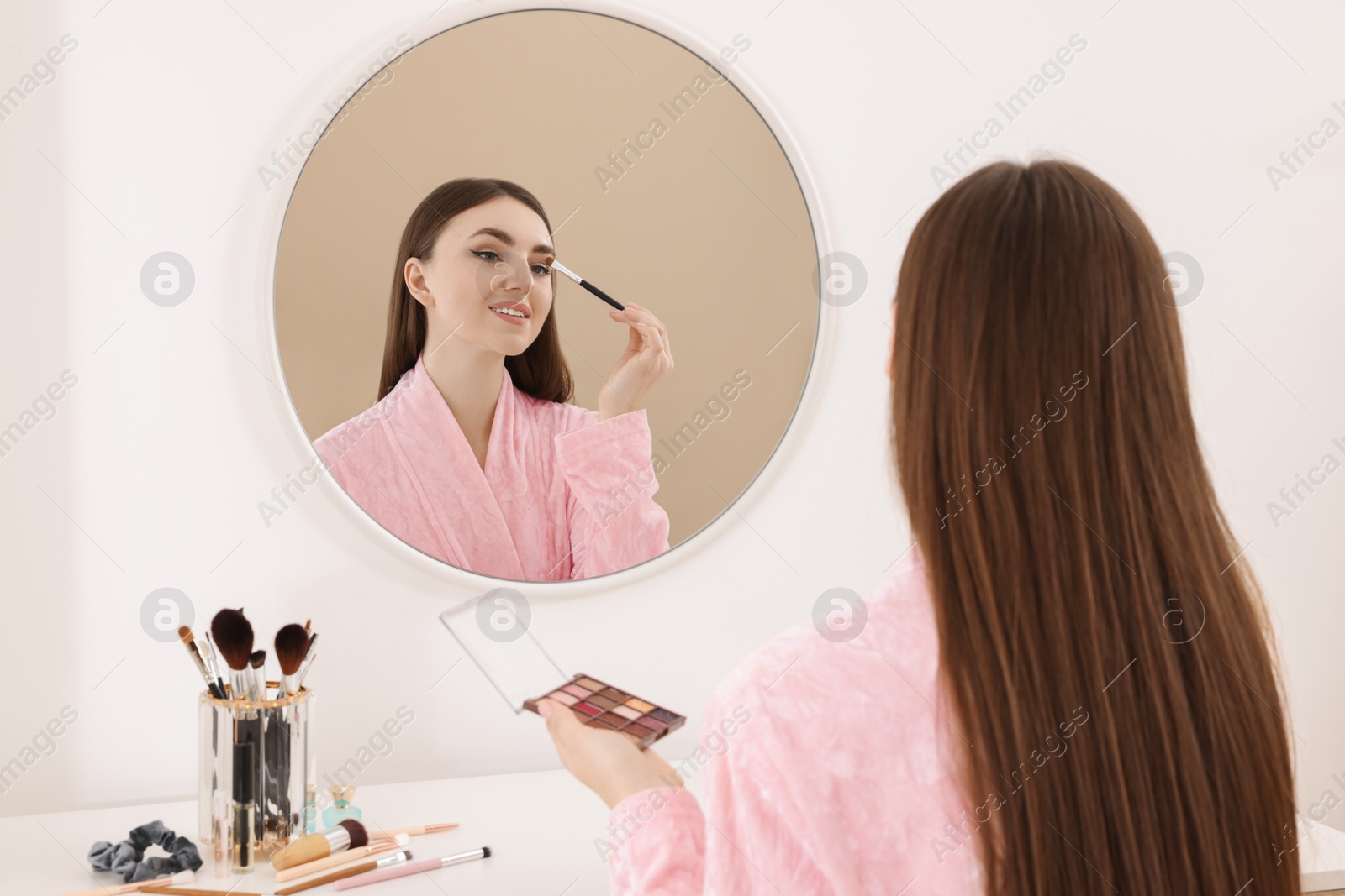Photo of Beautiful young woman applying eyeshadow with brush near mirror indoors