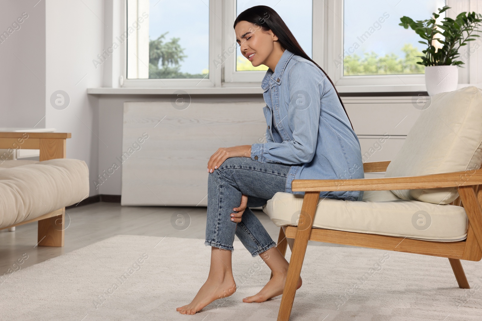 Photo of Woman suffering from leg pain on armchair at home