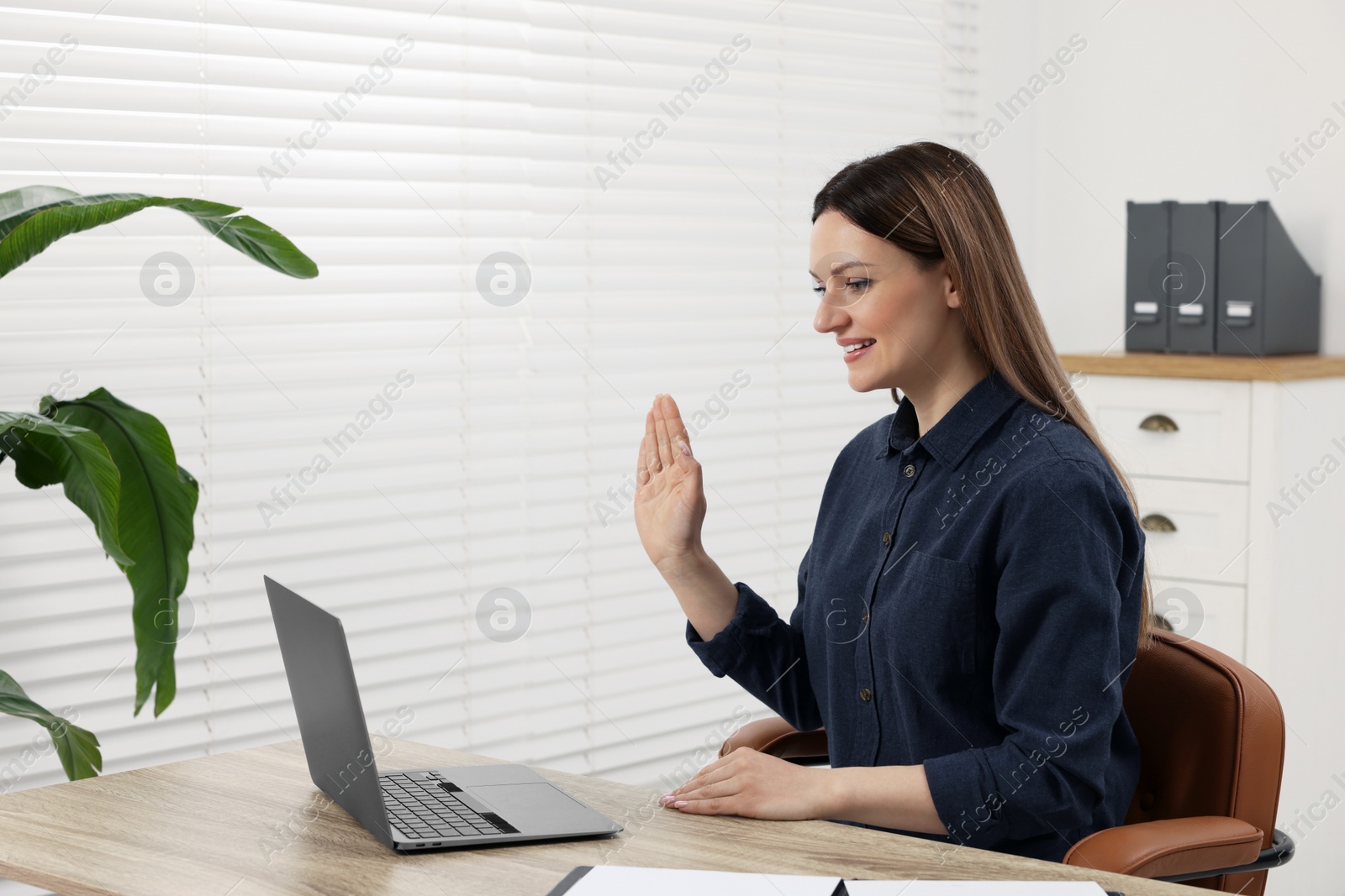 Photo of Woman waving hello during video chat via laptop at wooden table in office