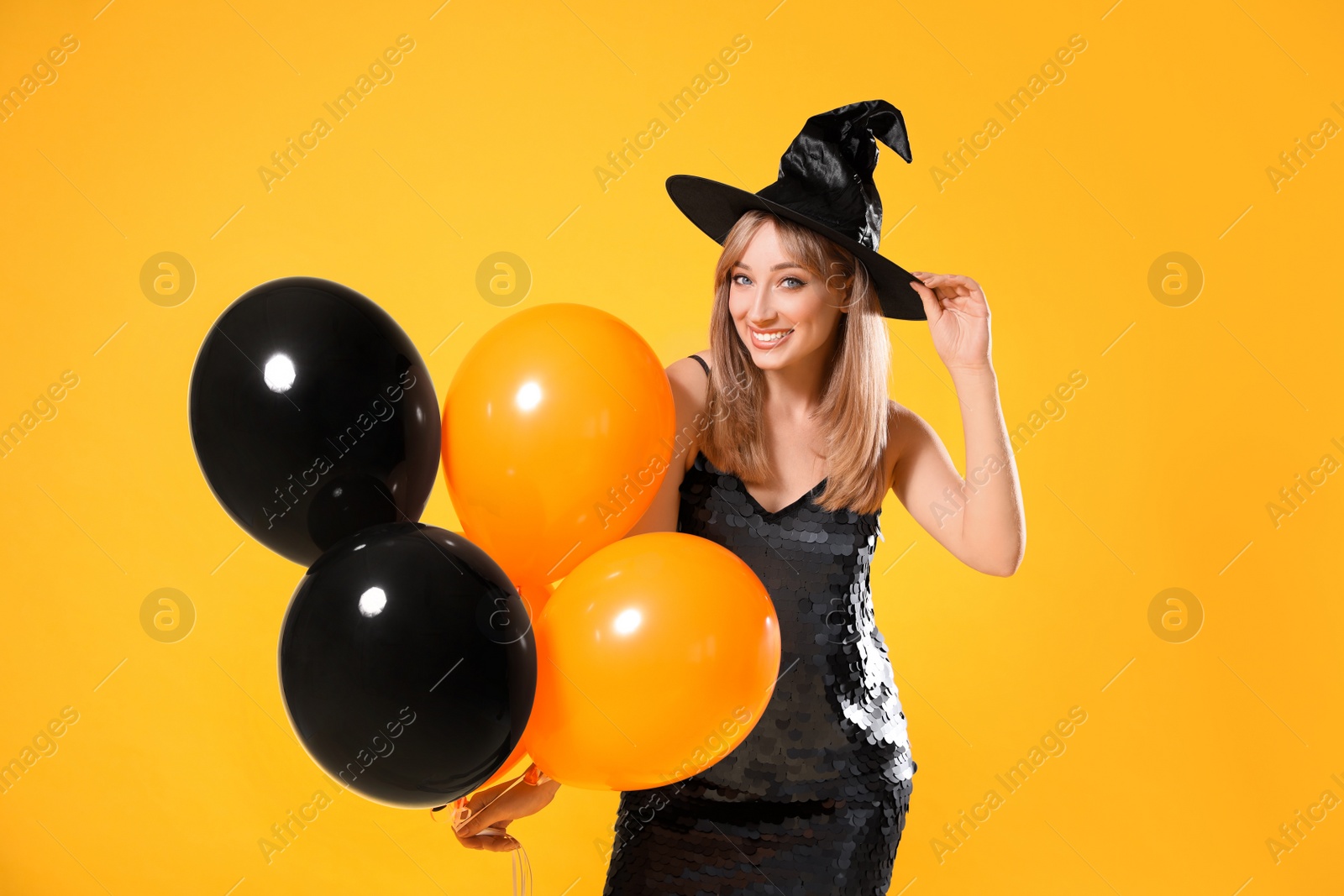 Photo of Beautiful woman in witch costume with balloons on yellow background. Halloween party