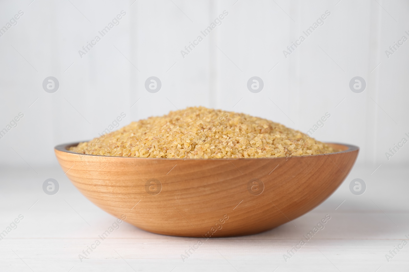 Photo of Bowl of uncooked bulgur on white wooden table, closeup