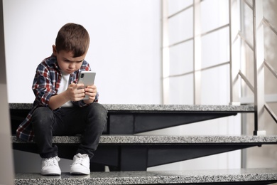 Sad little boy with mobile phone sitting on stairs indoors