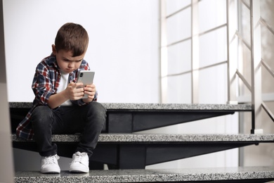 Photo of Sad little boy with mobile phone sitting on stairs indoors