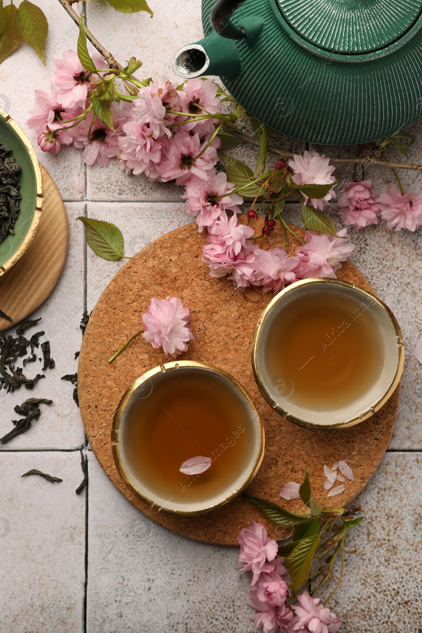 Photo of Traditional ceremony. Cup of brewed tea, teapot, dried leaves and sakura flowers on tiled table, flat lay