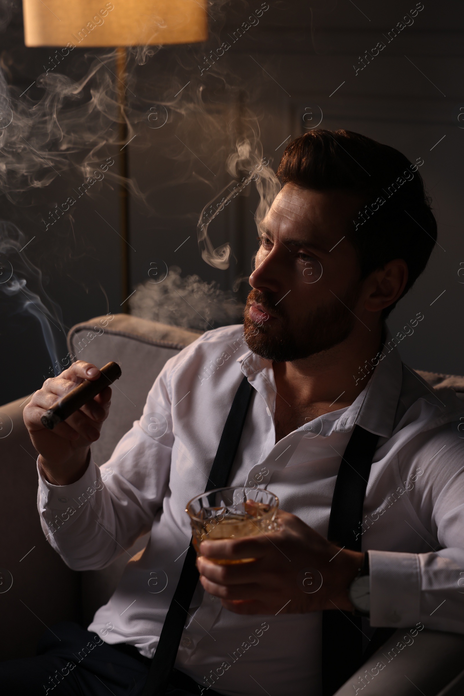 Photo of Handsome man with glass of whiskey smoking cigar in armchair at home