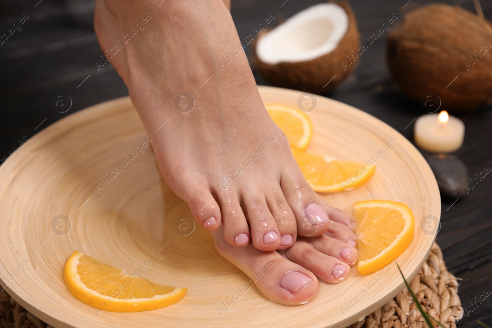 Photo of Woman soaking her feet in plate with water and orange slices on wooden floor, closeup. Spa treatment