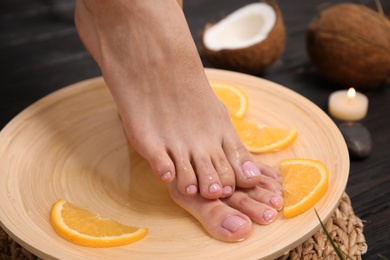 Woman soaking her feet in plate with water and orange slices on wooden floor, closeup. Spa treatment